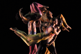 A cluster of dancers lifting and supporting eachother in front of a black background.